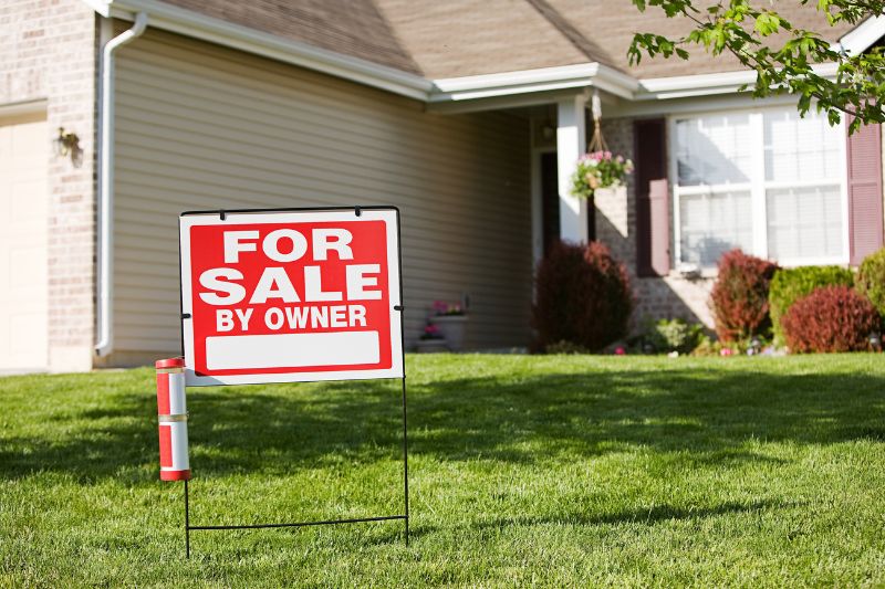 A For Sale by Owner sign is displayed on the front lawn of a suburban house with beige siding, white trim, and maroon shutters on a sunny day.