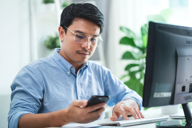 A man wearing glasses is sitting at a desk with computer and cell phone